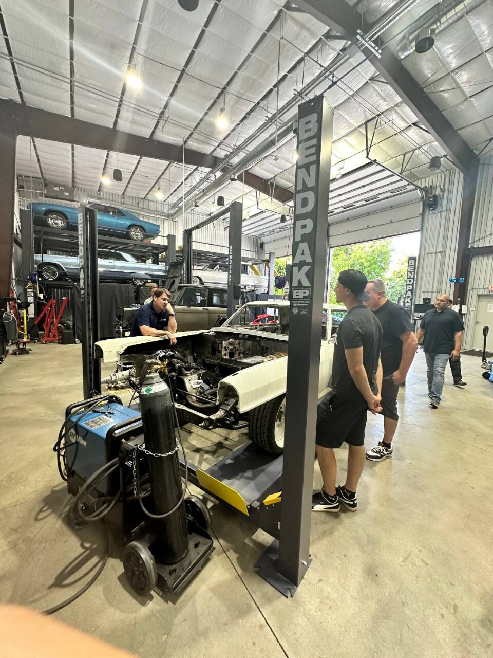 Jim Farley talks with the team at Current LA, an electric vehicle conversion shop focusing on vintage vehicles. They surround a vehicle currently being turned into an electric vehicle.